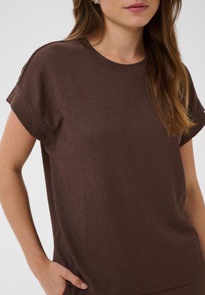 Femme aux longs cheveux bruns portant un haut marron texturé, ample et à manches courtes, une main dans la poche, sur un fond uni.