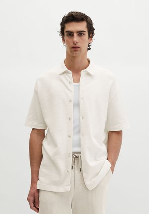 Junger Mann mit lockigem braunem Haar, trägt ein weißes kurzärmeliges Hemd mit Knöpfen über einem weißen Tanktop und beigen Hosen mit Kordelzug, stehend.