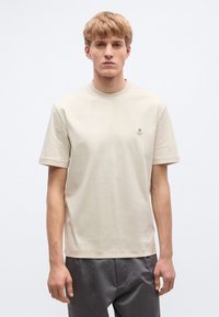 Μπεζ βαμβακερό μπλουζάκι με λαιμόκοψη και κοντά μανίκια, με μικρή στρογγυλή λεπτομέρεια λογότυπου στο αριστερό στήθος. Ομαλή υφή, άνετη γραμμή.