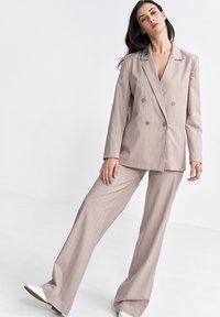 Beżowy garnitur w paski składający się z podwójnie zapiętej marynarki z szerokimi klapami oraz luźnych, szerokich spodni. Tkanina wydaje się gładka i dobrze ustrukturyzowana.