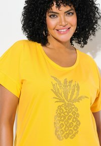 Camiseta amarilla de manga corta hecha de una tela suave, con un diseño brillante de piña plateada en la parte delantera, con un cuello redondo y un corte relajado.