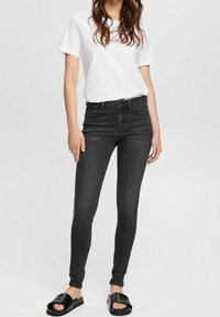 Vaqueros skinny negros de denim, de cintura alta con un diseño clásico de cinco bolsillos, combinados con una camiseta blanca y sandalias deslizantes negras.