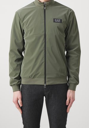 Homme portant une veste zippée vert olive avec le logo EA7 sur la poitrine et un pantalon foncé, debout devant un fond clair uni.
