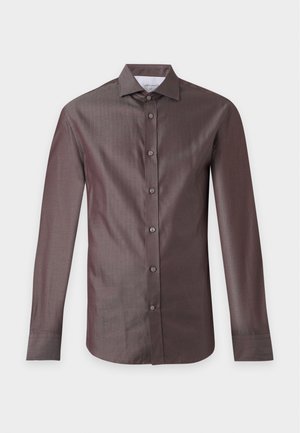 Man staat en draagt een lila overhemd met knopen, donkerblauwe broek en zwarte en beige loafers tegen een effen grijze achtergrond.