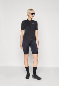 Svart cykeltröja med korta ärmar, hel dragkedja och logotyp; matchad med svarta vadderade shorts och svarta cykelskor. Bärande spegelglasögon.