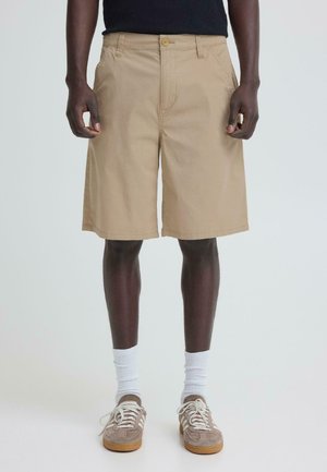 Uomo che indossa pantaloncini beige fino al ginocchio, calzini bianchi alla caviglia e scarpe da ginnastica beige con lacci bianchi, in piedi contro uno sfondo chiaro e uniforme.