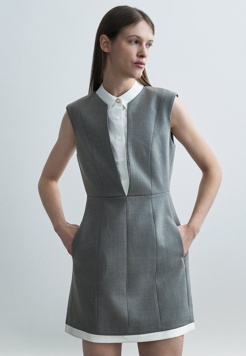 Γυναίκα με ίσια καστανά μαλλιά που φορά γκρι αμάνικο φόρεμα πάνω από λευκό πουκάμισο με κουμπιά, τα χέρια στις τσέπες, κοιτάζει προς τα δεξιά σε απλό φόντο.