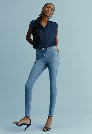 Marinblå ärmlös skjorta med knappp detaljer, matchad med ljusblå skinny jeans. Modellen bär svarta remsandaler mot en ljusblå bakgrund.
