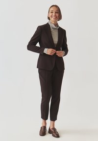 Smiling woman adjusting dark brown blazer over gray turtleneck, wearing matching pants and brown loafers, standing on light background.