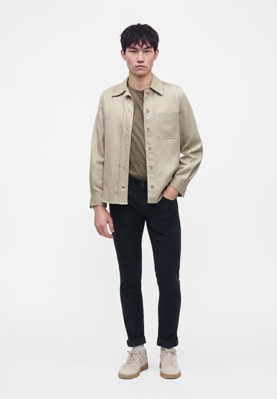 Jeune homme debout portant une veste beige à boutons, une chemise vert olive, un pantalon noir et des baskets blanches, une main dans la poche.