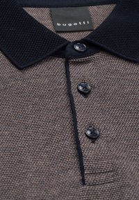 Polohemd aus strukturiertem braunem Stoff mit einem dunkelblauen Kragen. Es hat drei schwarze Knöpfe und ein Etikett mit der Aufschrift "bugatti".