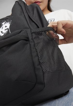 La main d'une personne insérant les doigts dans la poche latérale en maille noire d'un sac à dos noir avec un graphisme blanc sur la partie supérieure.