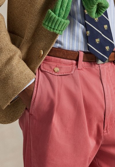 Ροζ παντελόνι από κορντούρα με εμπρόσθιες τσέπες, καφέ δερμάτινη ζώνη και γραβάτα με σχέδιο. Καφέ υφασμάτινο σακάκι και πράσινο πλεκτό κασκόλ εμφανή.