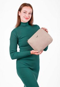 Béžová prošívaná kabelka s texturovan�ým povrchem a zipovým uzávěrem, držená osobou v zelených žebrovaných šatech s rolákem na bílém pozadí.