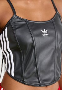Top cropped preto feito de couro sintético, com um design de três riscas brancas nas laterais e o logótipo da Adidas em branco bordado na parte da frente.