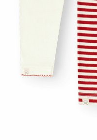 Manches longues de deux chemises pour bébé, l'une unie crème avec bordure rouge, l'autre rayée rouge et blanche, toutes deux avec de petites étiquettes de marque en tissu.
