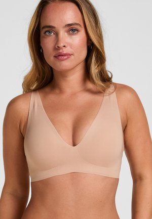 Woman with light skin and blonde hair wearing a nude-colored seamless bra with wide straps, facing forward against a plain light background.