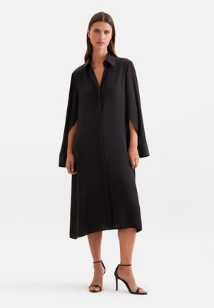 Femme portant une robe midi noire ample avec des manches style cape et des sandales noires à talons hauts, debout devant un fond uni.
