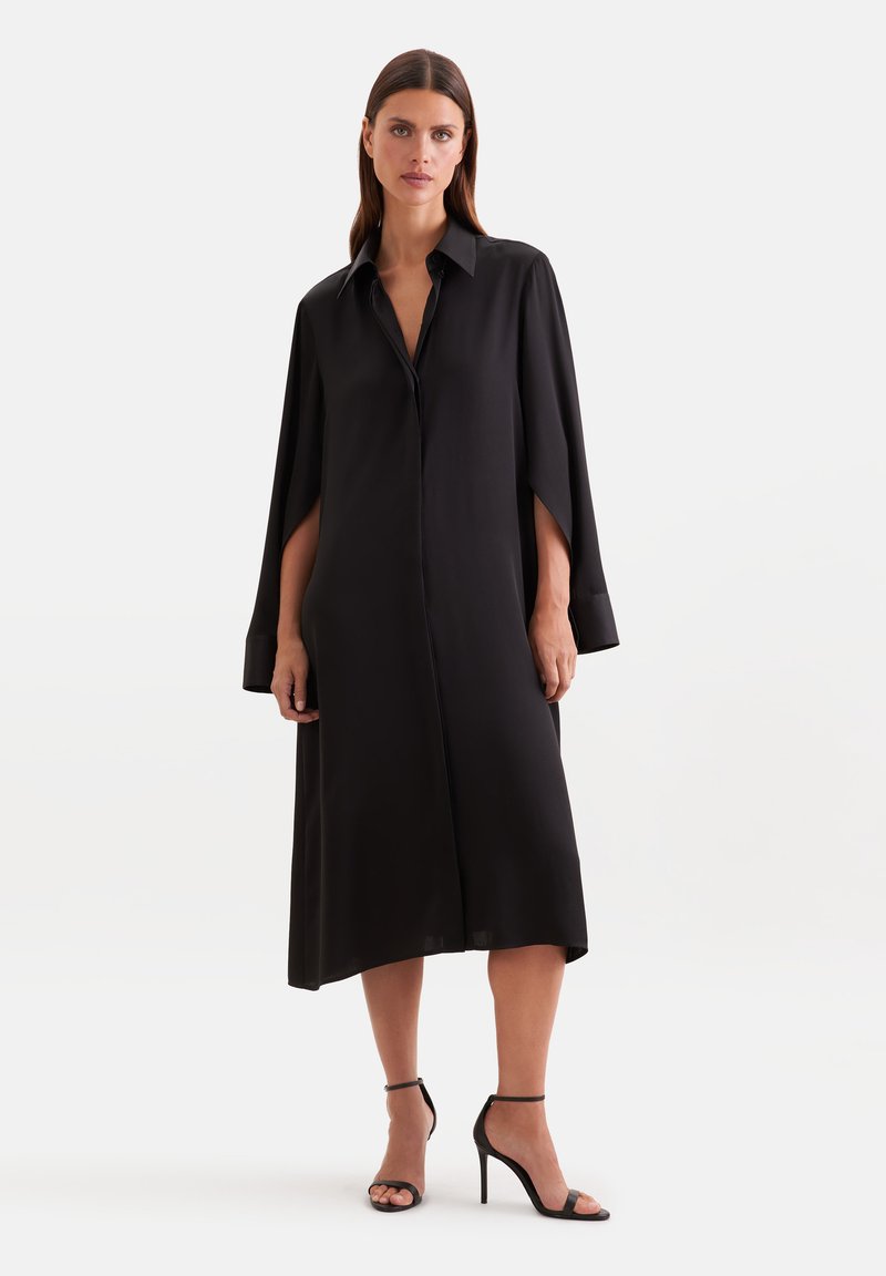 Femme portant une robe midi noire ample avec des manches style cape et des sandales noires à talons hauts, debout devant un fond uni.