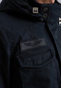 Giacca blu navy realizzata in tessuto resistente, con zip frontale, cappuccio, toppa sul petto con design di ali e tasca laterale.