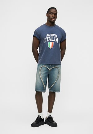 Blue graphic t-shirt with rolled short sleeves featuring white text and an Italian shield. Distressed denim shorts with heart-shaped tears. Black sneakers.