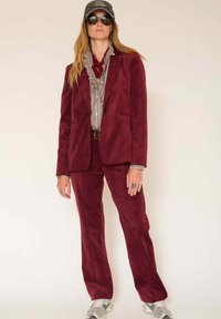 Costume en velours côtelé bordeaux avec un blazer ajusté et un pantalon droit. Superposé avec une chemise grise à volants et terminé avec des baskets décontractées.