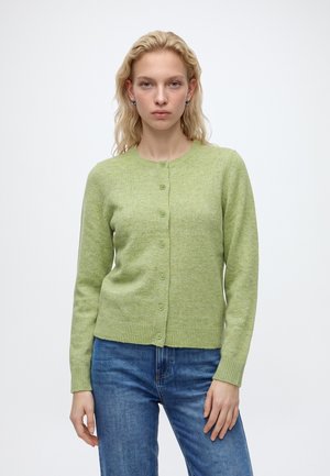 Jeune femme aux cheveux blonds portant un cardigan vert clair boutonné et un jean bleu, debout devant un fond clair uni.