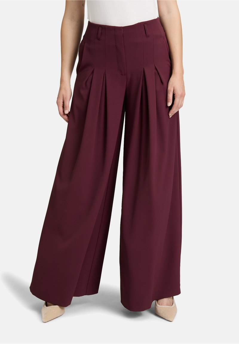 Pantaloni larghi borgogna con pieghe, realizzati in tessuto morbido. Presentano tasche laterali e una vita pulita, abbinati a tacchi di colore chiaro.