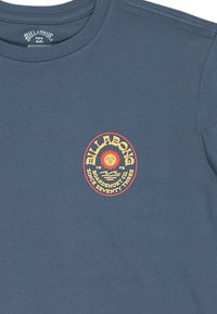 Modrá tričko s logom slnka v žltej a oranžovej farbe a textom "Billabong Boardshort Co. Od roku Sedemdesiat Tri" na hrudi.