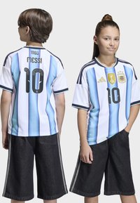 Dwoje dzieci w koszulkach reprezentacji Argentyny z numerem 10 i napisem "Messi" z tyłu; jedno z nich zwrócone przodem, drugie pokazuje tył koszulki.