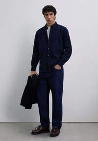 Camisa de botones navy con cuello, combinada con jeans oscuros y zapatos marrones de cordones. Textura textil ligera con una silueta ajustada.