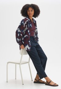Meerkleurig overhemd met abstract patroon, donkerpaarse tanktop, marineblauwe broek en bruine sandalen, zittend op een witte stoel.