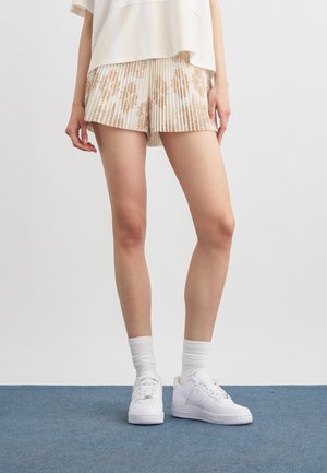 Person iført hvide plisserede shorts med brune blomstermønstre, hvide sneakers og hvide sokker, stående på blå tæppe.