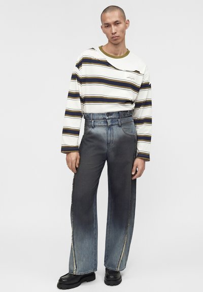 Άνδρας με λευκό ριγέ πουκάμισο μακριά μανίκια, μέσα σε ψηλόμεσο, φαρδύ ξεθωριασμένο τζιν και μαύρα παπούτσια, όρθιος μπροστά σε απλό φόντο.