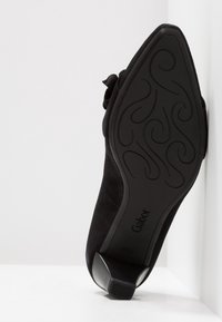 Chaussure à talon haut en daim noir avec un bout pointu et un nœud décoratif. Dotée d'une semelle en caoutchouc noir texturée et d'un talon large et bas.