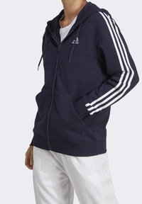 Person som bär en marinblå zip-up hoodie med vita ränder på ärmarna och vita byxor, står med en hand i fickan.