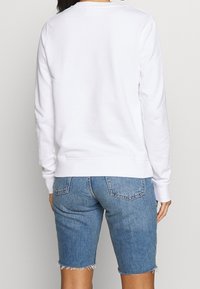 Bílá mikina s dlouhými rukávy a kulatým výstřihem, kombinovaná s modrými džínovými šortkami s frayed lemem. Minimalistický design, bez viditelných vzorů.