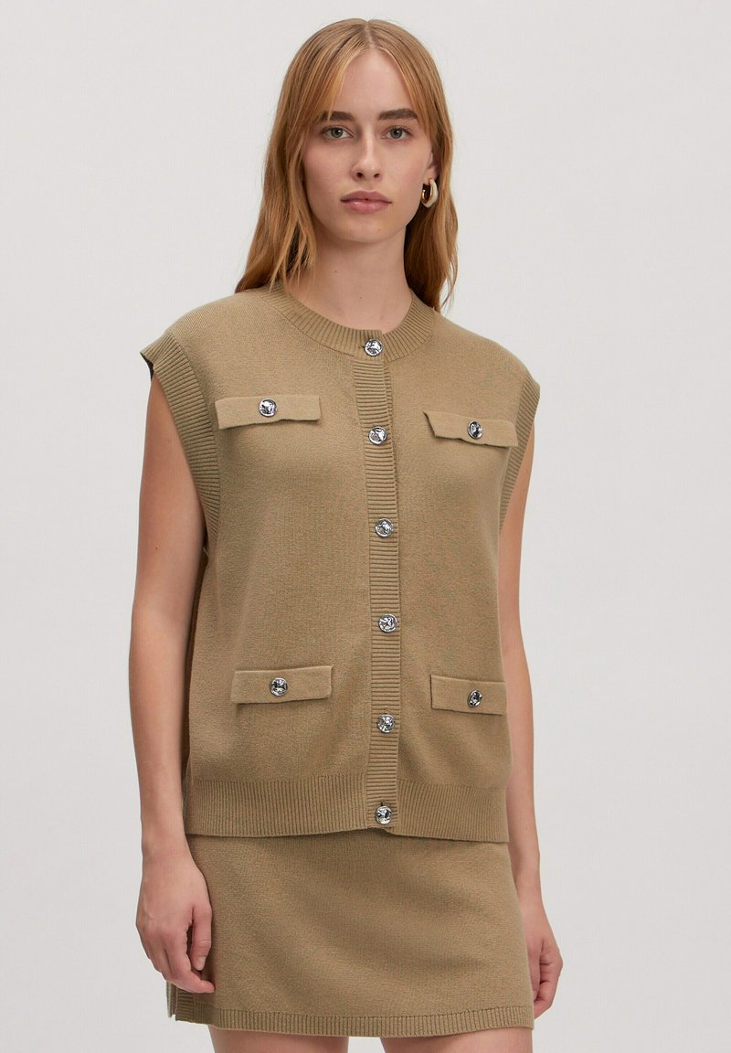 Khaki ärmellose Strickjacke mit geripptem Halsausschnitt, vorderer Knopfleiste und zwei Brusttaschen mit glänzenden silbernen Akzenten. Weiche Stricktextur.