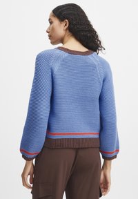 Blaue Strickjacke mit braunem Rippkragen und Saum, versehen mit roten und blauen Streifen am unteren Ende. Lange, gepuffte Ärmel.