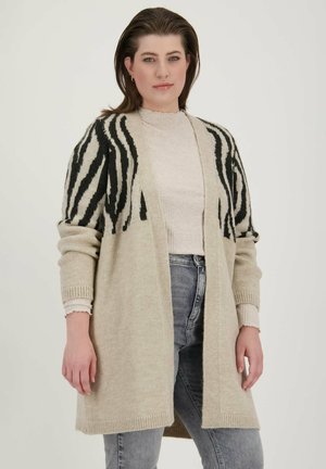 Pleteni kardigan v svetlo bež barvi z črnimi zebra črtami, odprt spredaj in z rebrastimi manšetami. Obučen preko teksturirane svetle majice in jeansa.