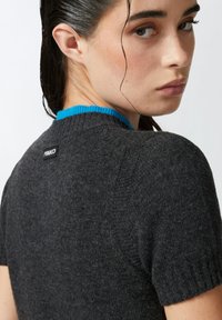 Pulover kratkih rukava u tamnosivoj pletenini s rebrastim ovratnikom u svijetloplavoj boji. Ima oznaku logotipa na stražnjoj strani izreza za vrat.