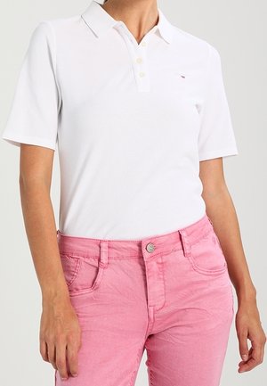 Biała koszulka polo z krótkim rękawem wpuszczona w wysokie różowe dżinsy na stojącej osobie, ręce swobodnie opuszczone wzdłuż ciała.