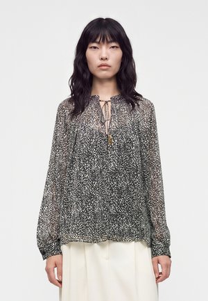 Femme aux longs cheveux noirs portant un chemisier transparent noir et blanc à motifs avec des manches longues et un lien au niveau du col, associé à une jupe blanche.