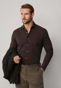 Hombre con camisa de botones marrón oscuro y pantalones marrón claro sostiene una chaqueta a cuadros oscuros con una mano en el bolsillo, mirando hacia un lado.