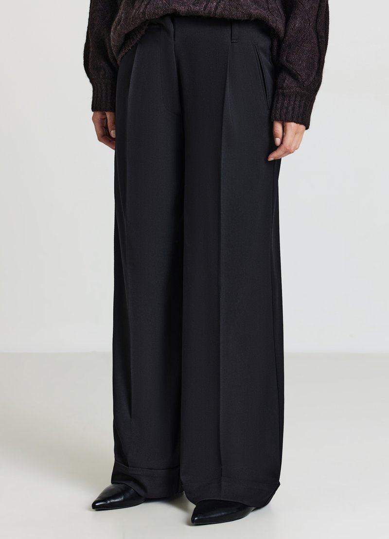 Schwarze weit geschnittene Hose aus glattem Stoff mit vorderen Falten, die eine taillierte Passform und eine weiche, fließende Textur aufweist.