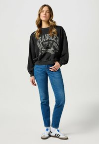 Czarna, oversize bluza z napisem i grafiką "Wrangler Motorcycle Club"; zestawiona z niebieskimi dżinsami i białymi trampkami z czarnymi akcentami.
