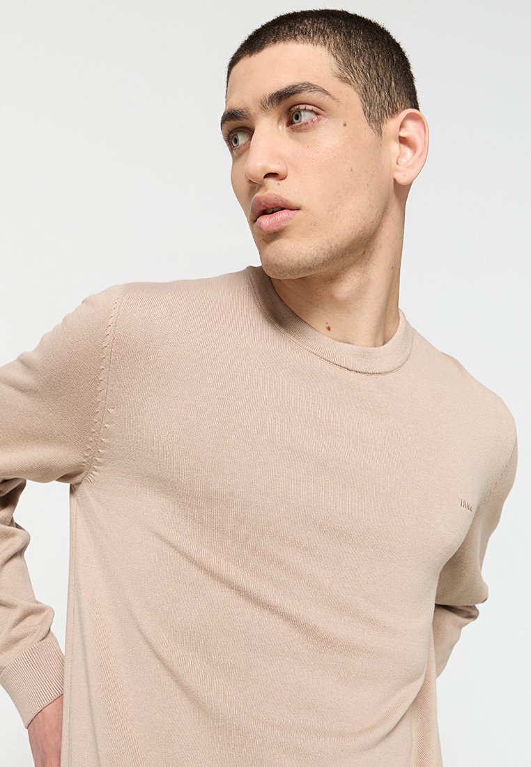 Beige gebreide trui met lange mouwen en een ronde halslijn, voorzien van subtiele textuur en gestikte details langs de schouder.