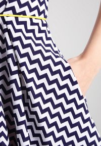 Main dans la poche d'une robe à motif chevron bleu marine et blanc, avec une fine ceinture jaune.