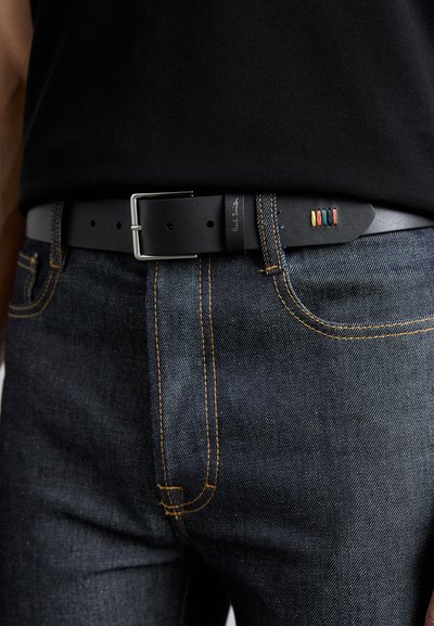 Svart lærbelte med en sølvmetallisk spenne, preget av en teksturert overflate og subtile fargeaksenter på enden, båret over denimjeans.