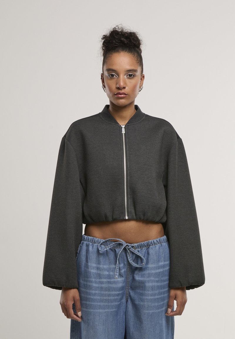 Femme aux cheveux bouclés en chignon portant une veste courte zippée gris foncé et un jean bleu ample à cordon, debout devant un fond uni.